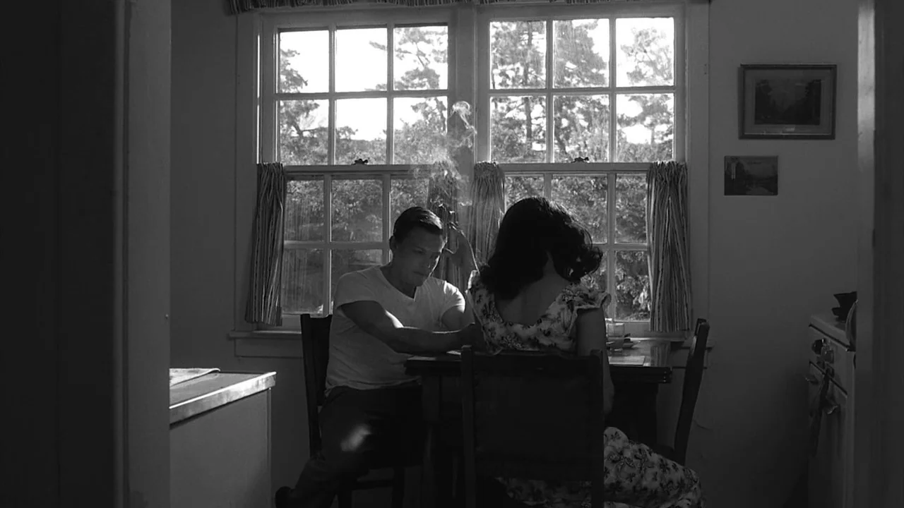 The Notorious Bettie Page (2005) movie still: medium — A small dining area with a window looking out onto trees. A man and a woman sit facing each…