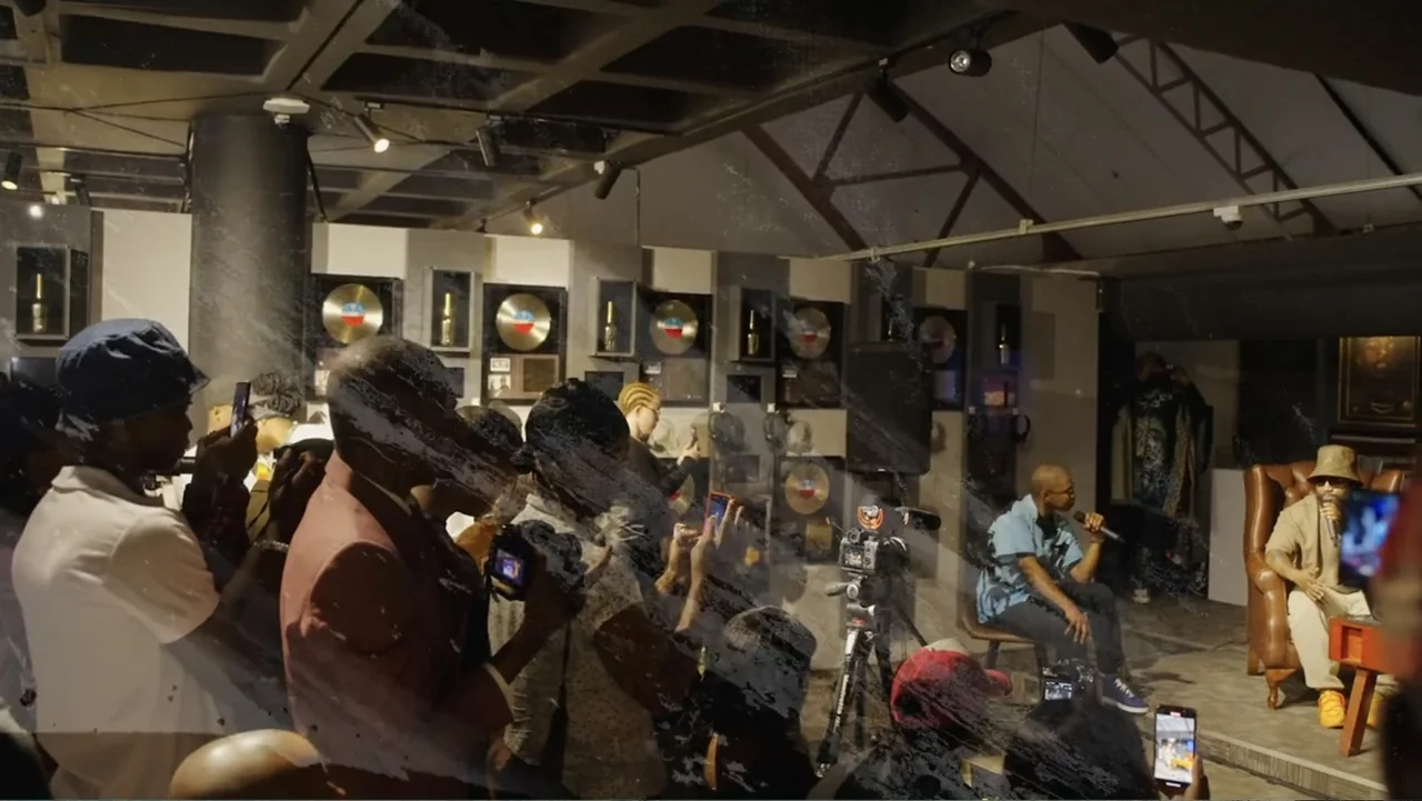 African Throne World Tour (2024) movie still: wide — A room filled with framed gold records on the wall and various onlookers holding phones. One…