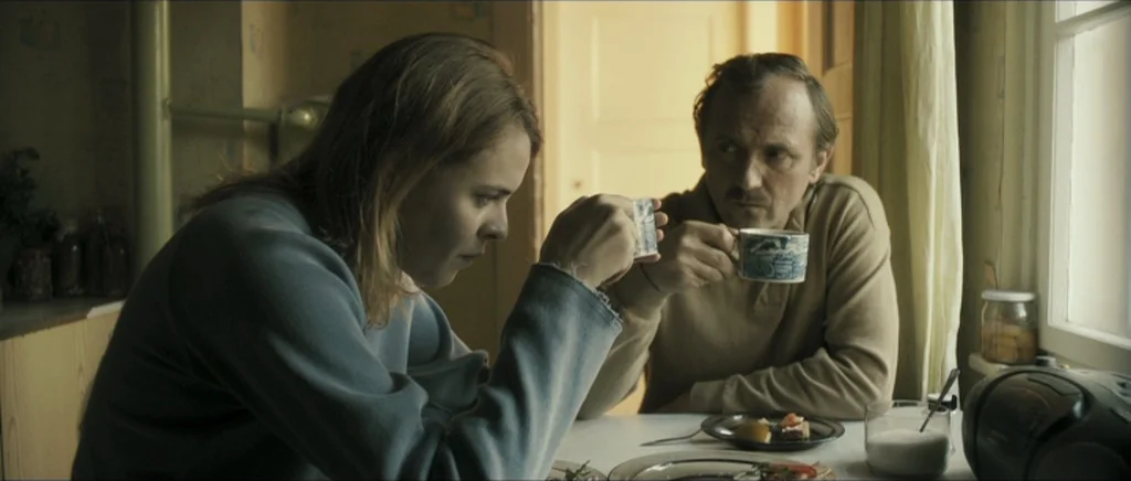 Lasting (Nieulotne) (2013) movie still: medium — A modest domestic interior with pale yellow wallpaper and a simple white table. A woman and a man…