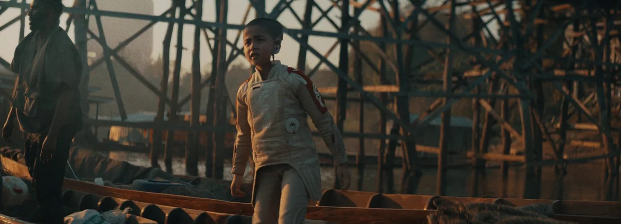 The Creator (2023) movie still: medium — A wooden dock structure with vertical support beams stands over water during sunset. A child in futuristic…
