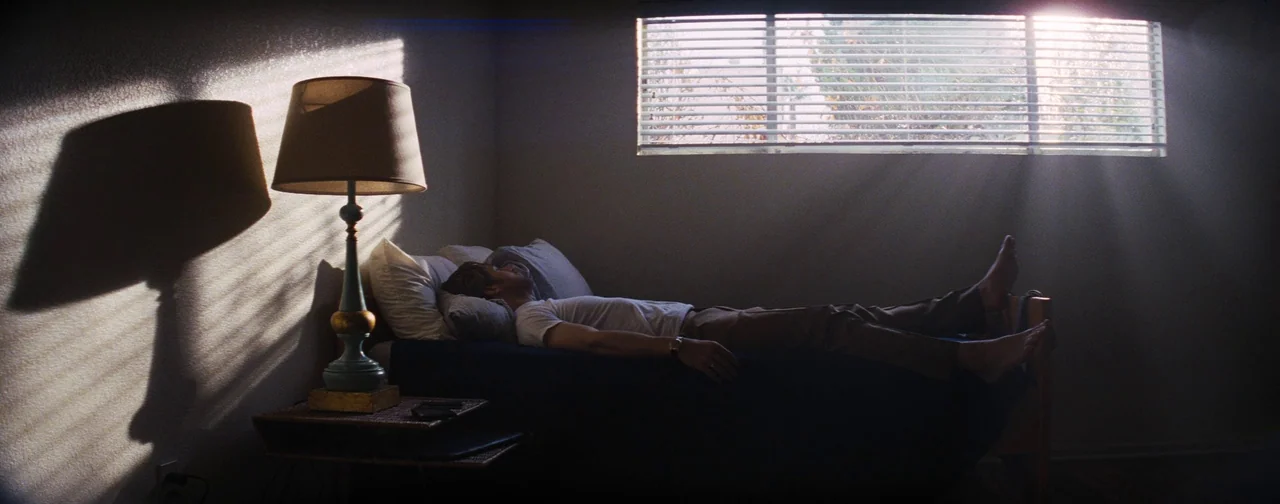 La La Land (2016) movie still: wide — A sparsely furnished room contains a bed and a bedside lamp with a shade. A man rests motionless on the bed…