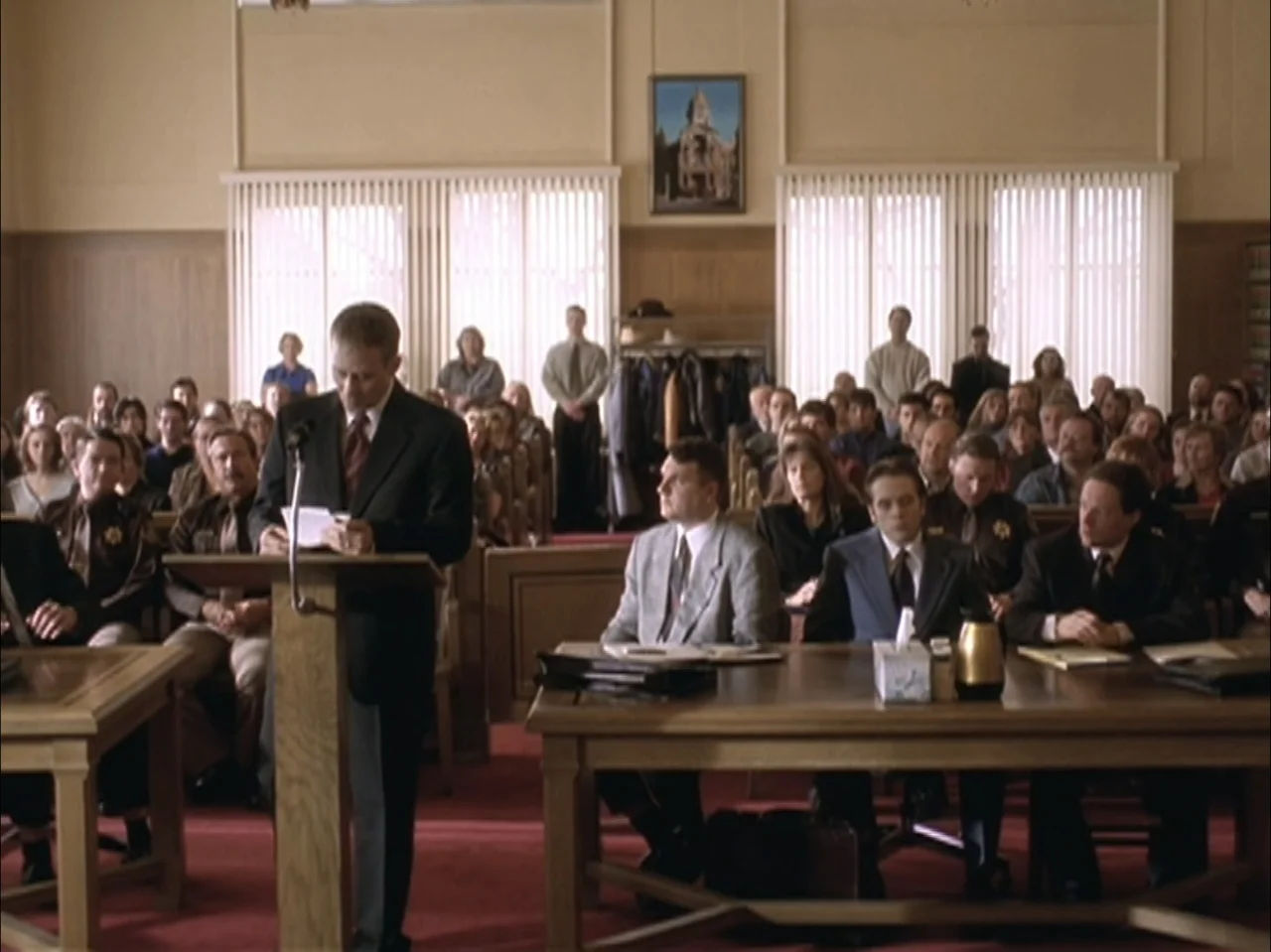 The Laramie Project (2020) movie still: wide — A wood-paneled room features rows of seated people facing a wooden lectern. A man stands at the…