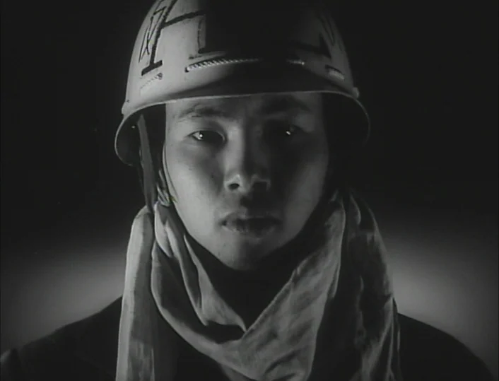 Funeral Parade of Roses (1969) movie still: closeup — A man wearing a military helmet and cloth neck wrap poses against darkness. The subject faces…