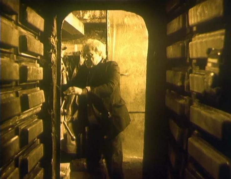 Dead Man’s Letters (1986) movie still: medium — A narrow passage is flanked by rows of stacked rectangular storage crates or drawers. A man in a…