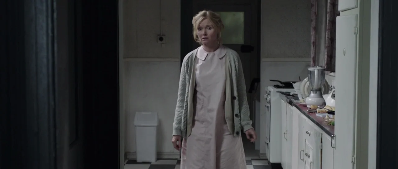 The Babadook (2014) movie still: fullbody — A 1950s kitchen features checkerboard floor tiles and white cabinets with a cluttered counter. A woman…