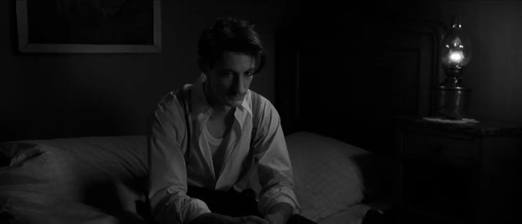 Frantz (2016) movie still: medium — A dimly lit space features a plain bed and a simple wooden bedside table. A young man sits on the edge of the…