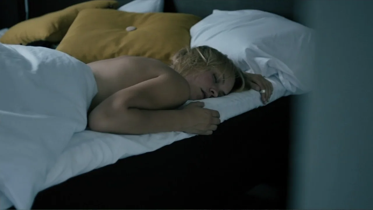 Copenhagen (2002) movie still: medium — A dark bedroom with white linens and mustard yellow decorative cushions on the bed. A blonde woman lies on…