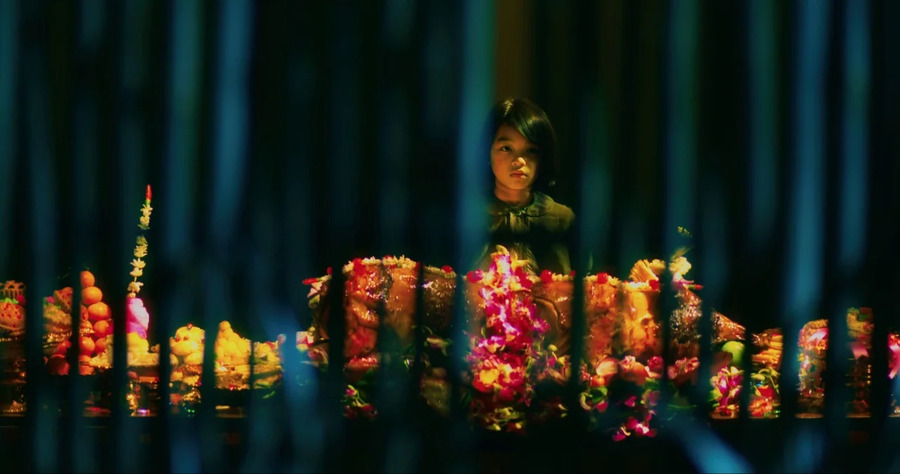 First They Killed My Father (2017) movie still: medium — A young girl stands behind a wooden table covered in ceremonial offerings and flowers.…