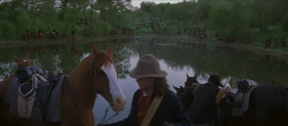 Ride With The Devil movie still: wide — A group of men on horseback pause near a quiet pond surrounded by trees. The cavalrymen are scattered along…