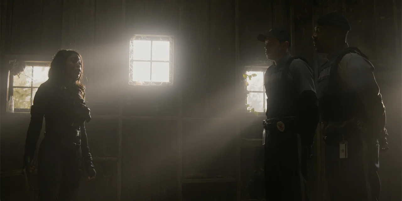 The Pickup (2025) movie still: medium — A dark, rustic interior features wooden walls and small windows with dust motes dancing. A woman in tactical…