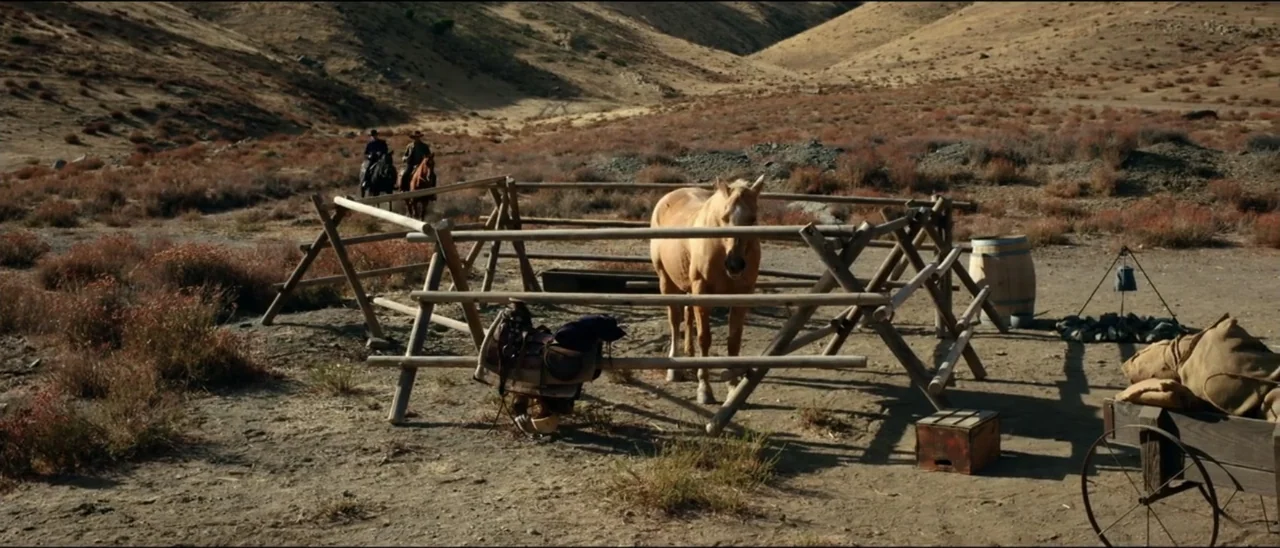 Dead for a Dollar (2022) movie still: wide — An arid, rocky canyon setting features a wooden corral in the foreground. Two men on horseback approach…