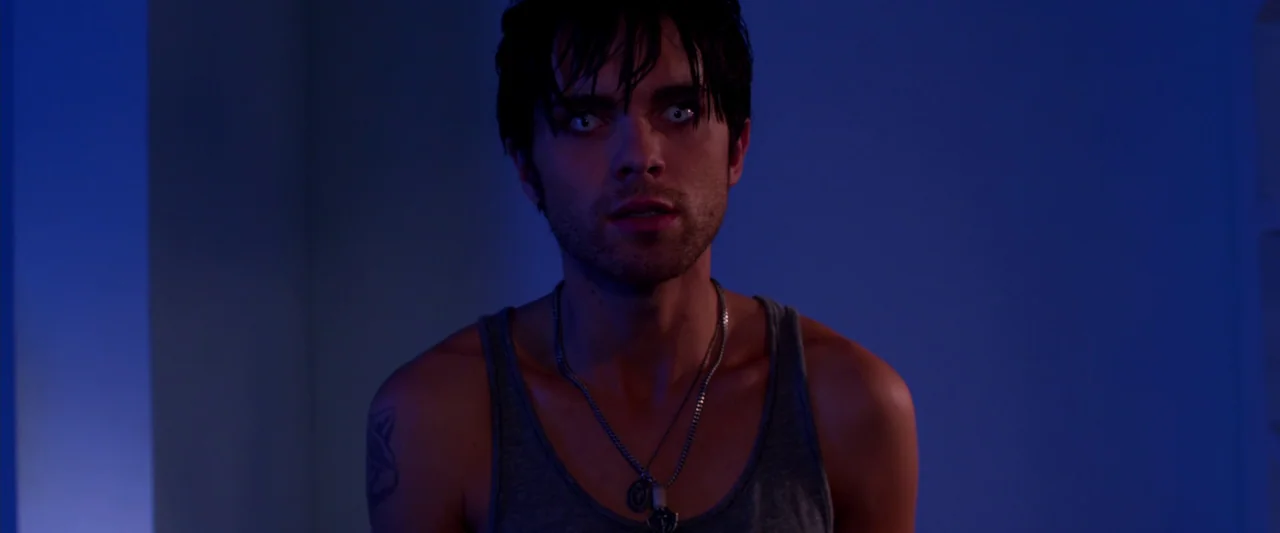 Kaboom (2010) movie still: medium — A dark indoor space is lit by a dim cool blue light source. A young man with white eyes stands centered in the…