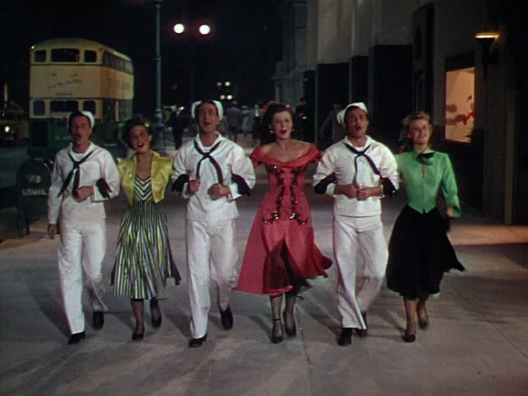On The Town (1949) movie still: fullbody — A group of six people walk across a concrete city sidewalk at night. A yellow double-decker bus is parked…