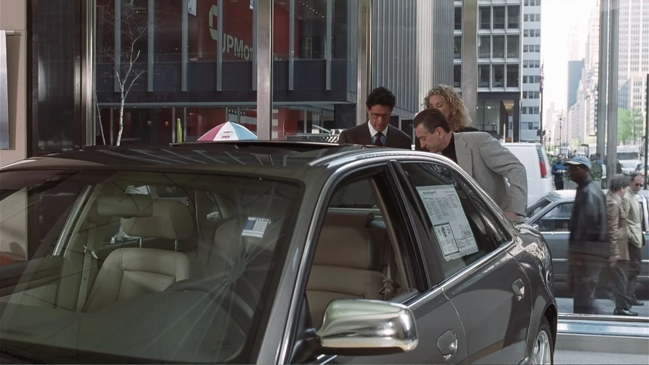 Analyze That (2002) movie still: medium — A silver sedan sits inside a glass-walled showroom with city buildings visible outdoors. A man inspects…