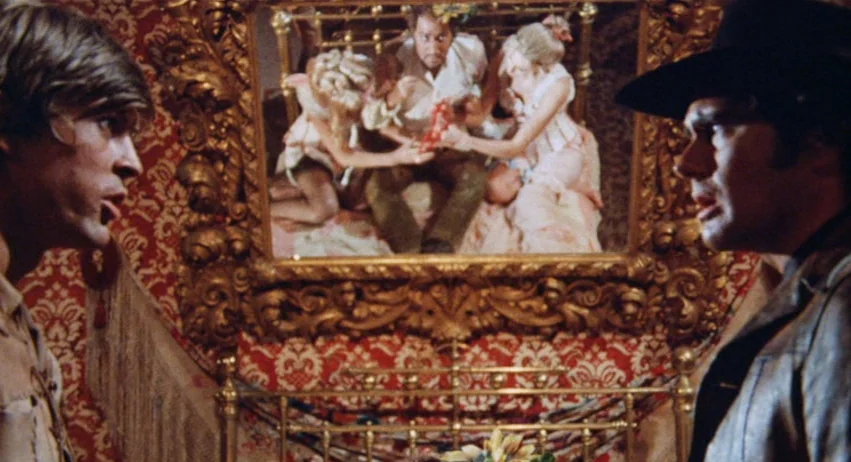 Roy Colt and Winchester Jack (1970) movie still: medium — A dimly lit room features ornate red patterned wallpaper and a large golden mirror. Two…