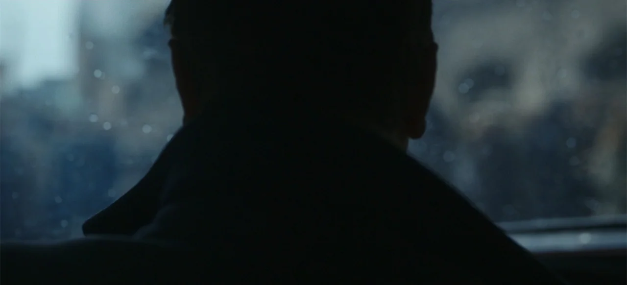 One Life (2023) movie still: closeup — A dark vehicle interior with water droplets visible on a rainy exterior window pane. A man sits facing the…
