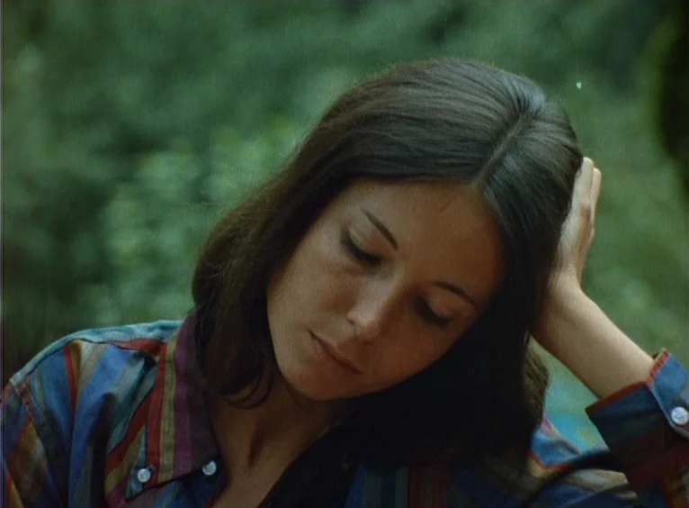 La Collectionneuse (1967) movie still: closeup — A woman sits in a leafy garden with soft sunlight filtering through the foliage. She rests her head…
