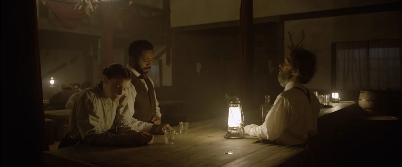 Ghosts of the Ozarks (2022) movie still: medium — A dimly lit 19th-century saloon features wooden walls and warm lantern light on the counter. A man…