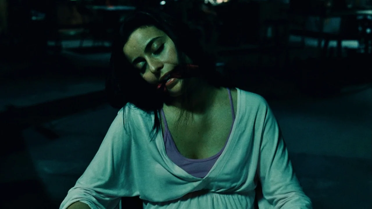 Saw III (2006) movie still: medium — A dimly lit room with cool-toned shadows surrounding the seated figure. A woman with dark hair sits with her…