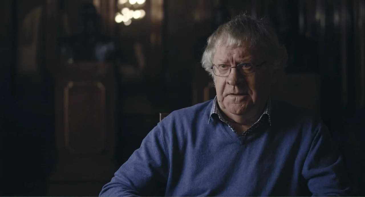 Los días azules (2025) movie still: medium — A dimly lit interior space with dark walls and an out-of-focus background light source. An older man…
