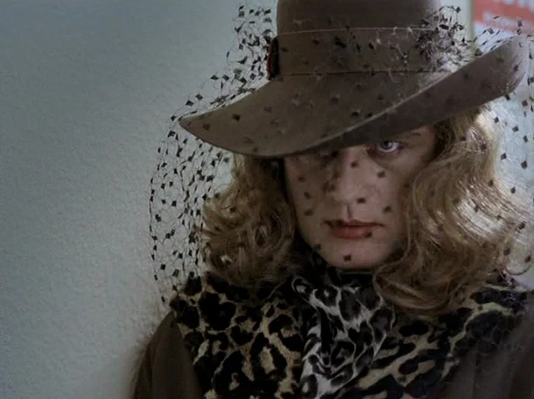 The Third Generation (1979) movie still: closeup — A person stands against a neutral off-white wall in an interior space. The subject gazes at the…