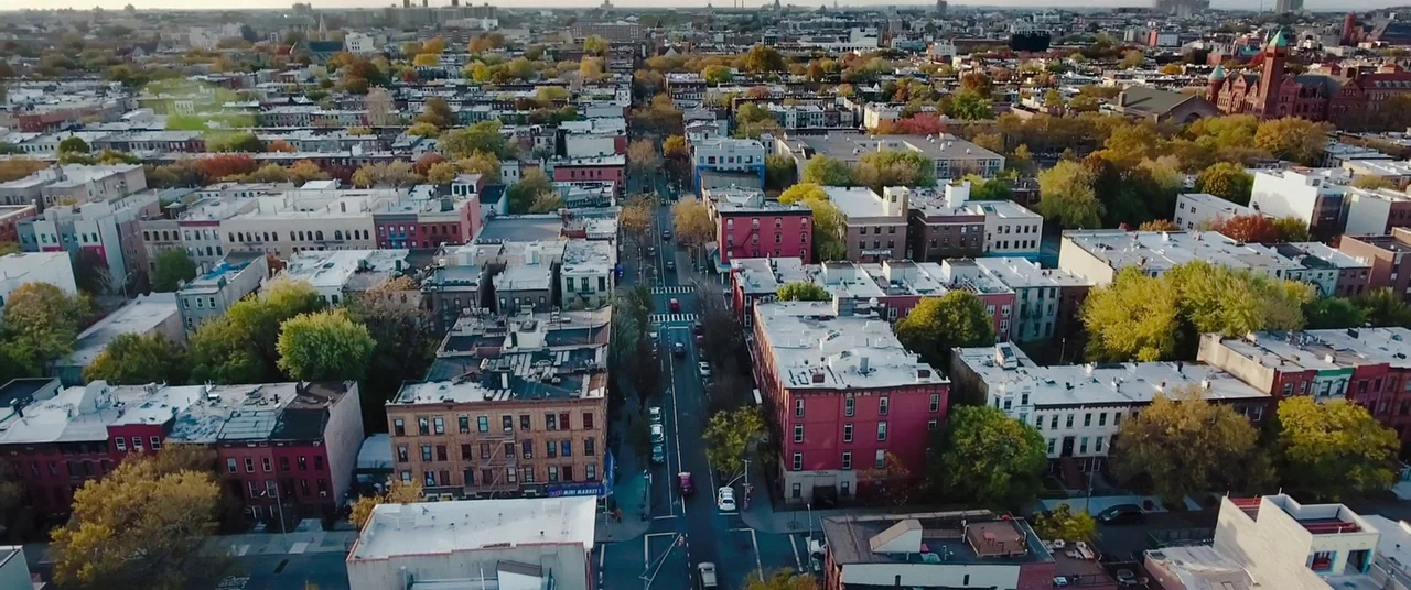 Child's Play (2019) movie still: establishing — A dense urban residential neighborhood features low-rise brick buildings and mature deciduous trees.…