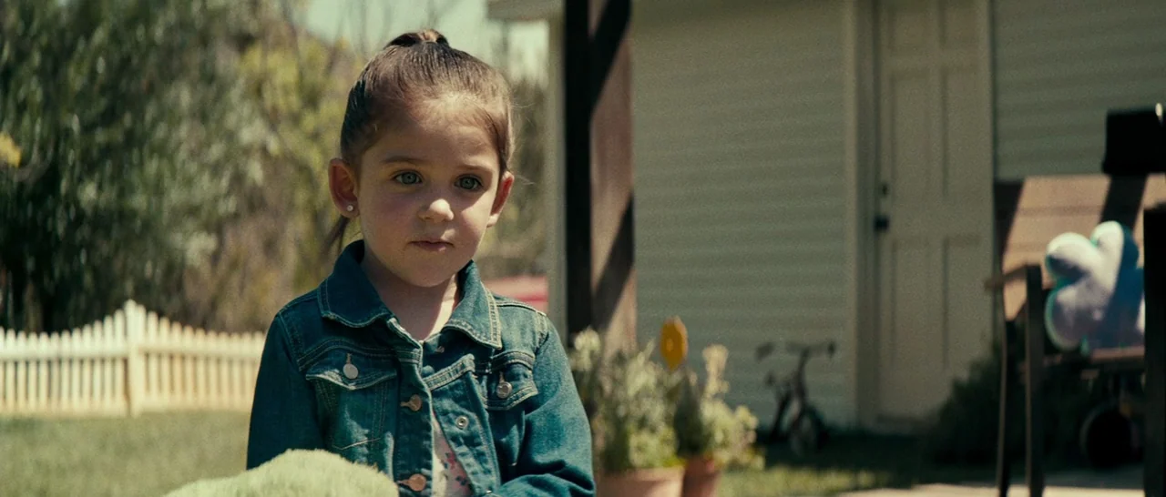 Swiss Army Man (2016) movie still: medium — A young child stands in a suburban front yard near a white house. She looks forward while wearing a…