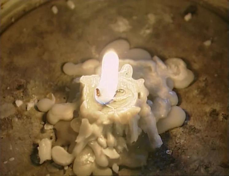 In The Presence of a Clown (1997) movie still: closeup — A close-up view of a partially melted white wax candle resting on a surface. The lit wick…