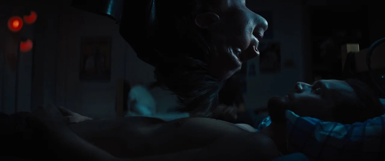 Daniel Isn’t Real movie still: medium — A dimly lit room with a person suspended upside down over a bed. A shirtless man lies on the pillow facing…