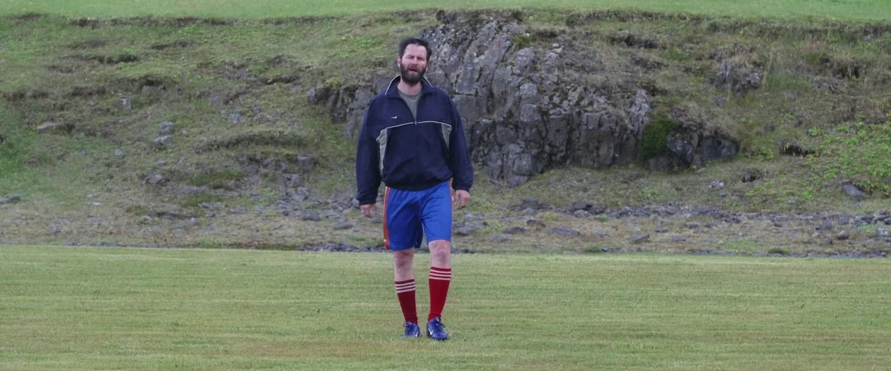 A White, White Day (2019) movie still: fullbody — An open grassy field sits in front of a rocky, uneven dirt embankment. A man in athletic gear…