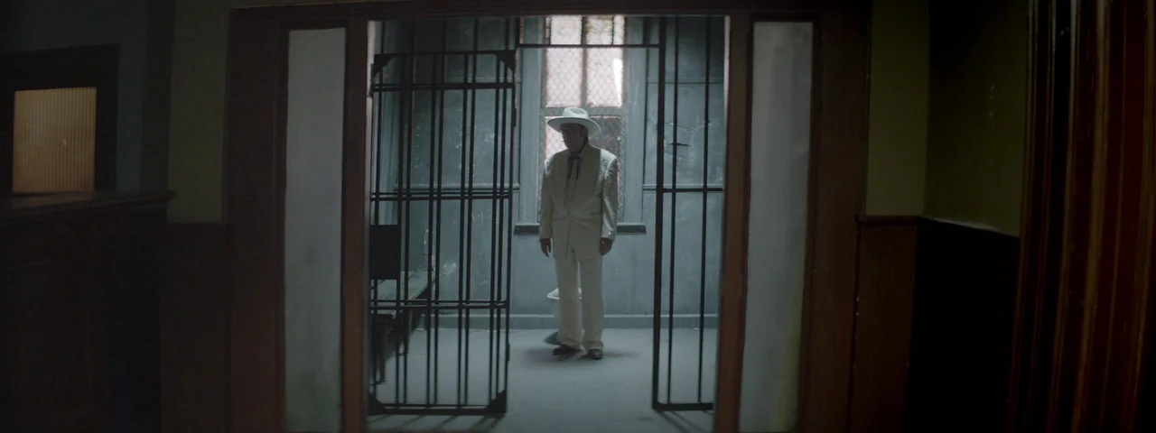 Entertainment (2015) movie still: fullbody — A dimly lit room looks into a barred space with a single toilet inside. An elderly man wearing a white…