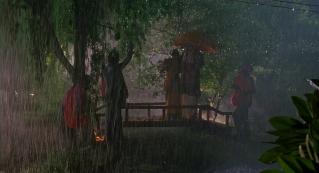 Monsoon Wedding (2001) movie still: wide — A wooden footbridge is surrounded by dense trees during a heavy rain shower. Multiple figures in…