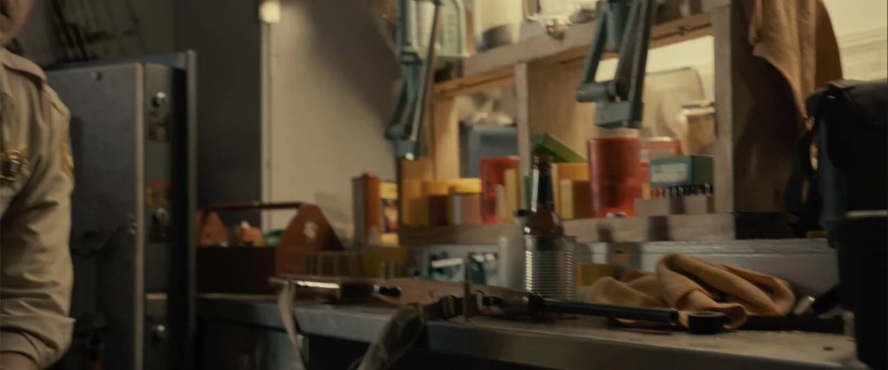 Frontera (2014) movie still: medium — A cluttered workshop workbench holds rifle parts, cleaning supplies, and a beer bottle. A person in a tan…