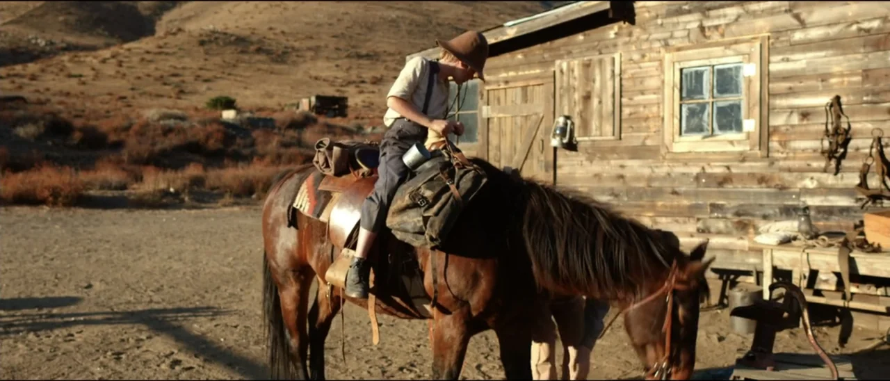 Dead for a Dollar (2022) movie still: medium — An arid ranch yard features a wooden structure beside a sun-drenched dusty clearing. A child wearing…