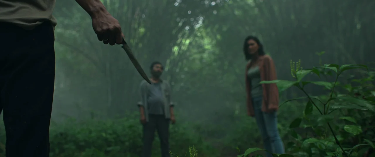 Impetigore (2019) movie still: medium — Two people stand in a dense, misty thicket of tall bamboo and green foliage. An unseen individual in the…