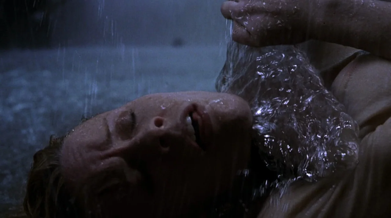 Hollow Man (2000) movie still: closeup — A person lies on the ground outdoors during a heavy rainstorm at night. Water falls directly onto the…