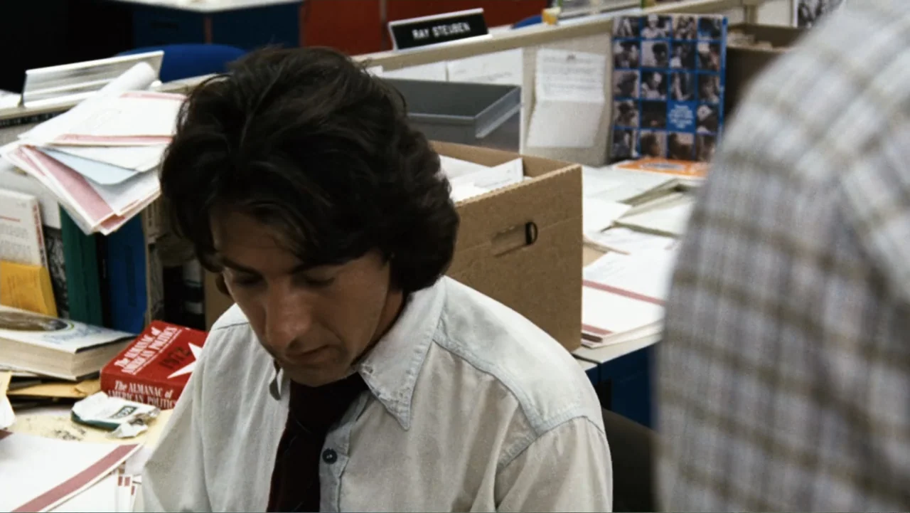 All the President's Men (1976) movie still: medium — A cluttered newspaper office desk is covered with stacks of paper and reference books. A man…