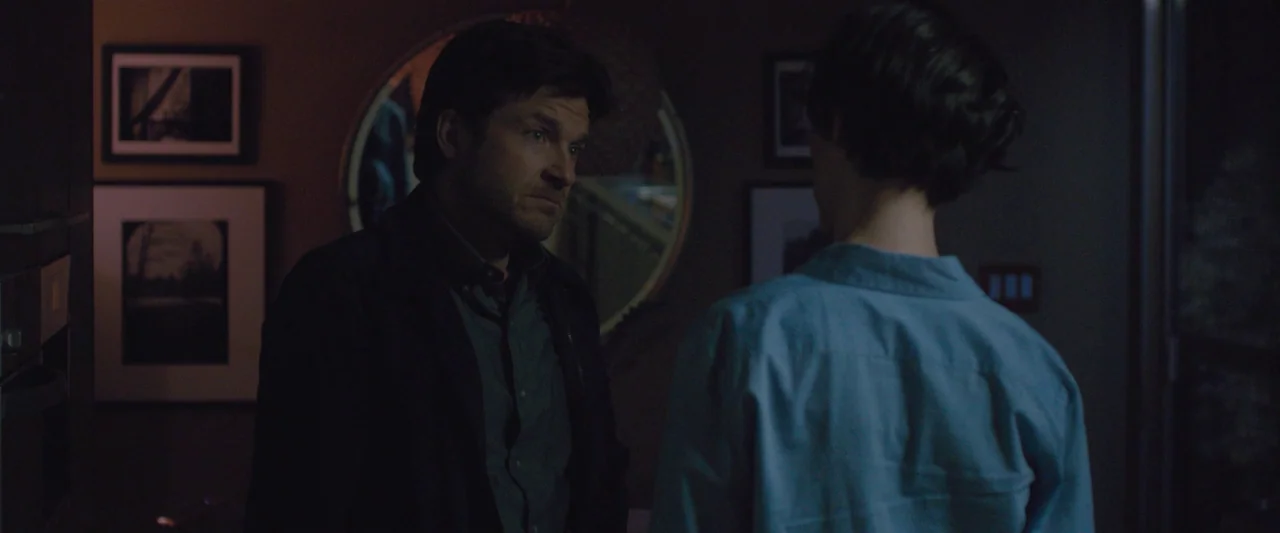 The Gift (2015) movie still: medium — A dark, residential interior hallway with framed art on the walls and a mirror. A man stands in profile…