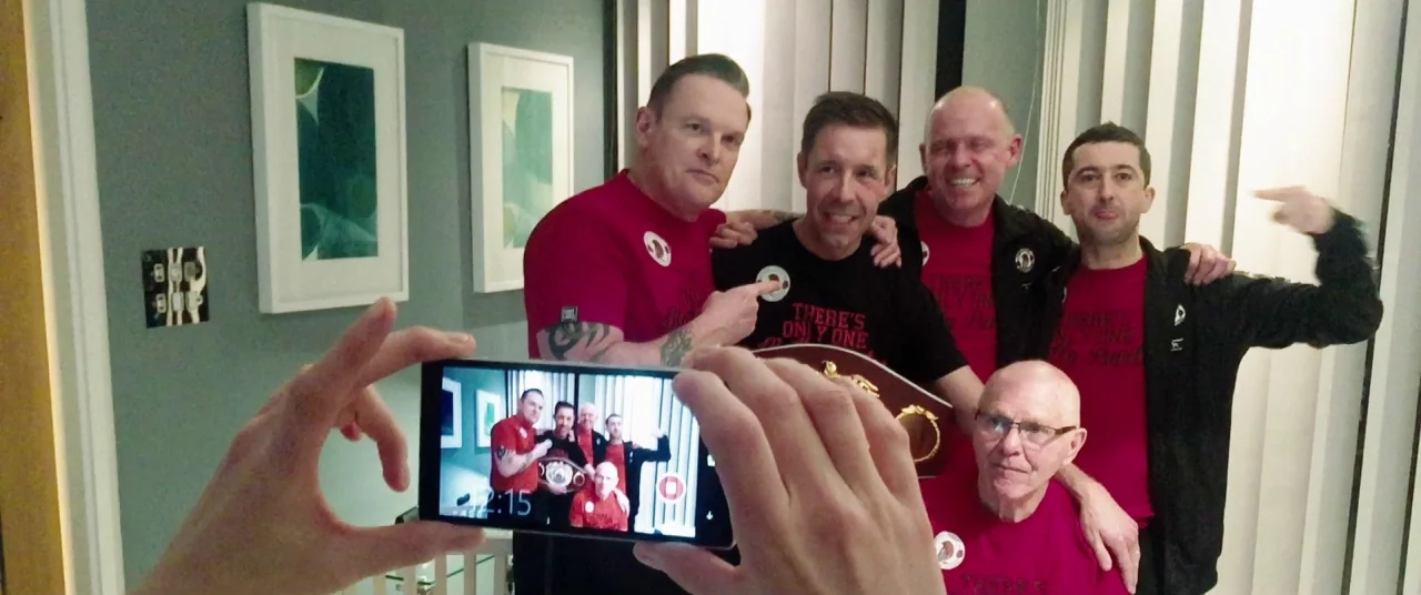 Journeyman (2017) movie still: medium — A group of five men in red matching t-shirts pose for a smartphone photograph. The men stand in front of a…