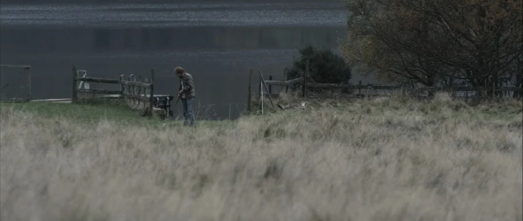 One Hundred Mornings (2009) movie still: wide — A vast expanse of dry, tall grass leads to a dark, still lake. A man stands beside a weathered…