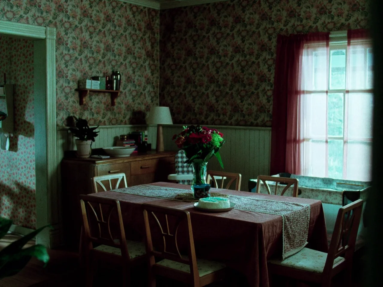 Die My Love (2025) movie still: wide — A vintage dining room features floral wallpaper above beige wainscoting and a wooden table. A single white…