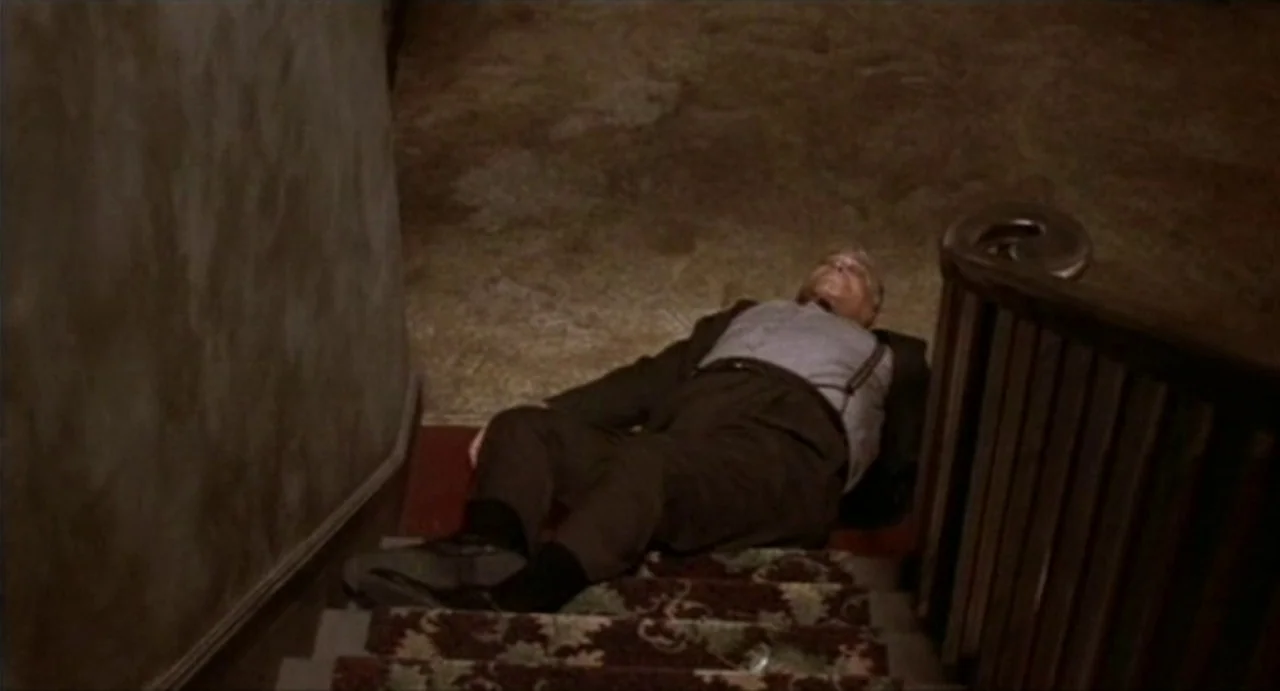The Annihilation of Fish (1999) movie still: fullbody — A dark, carpeted staircase with a wooden railing and a beige wall. An elderly man in a suit…