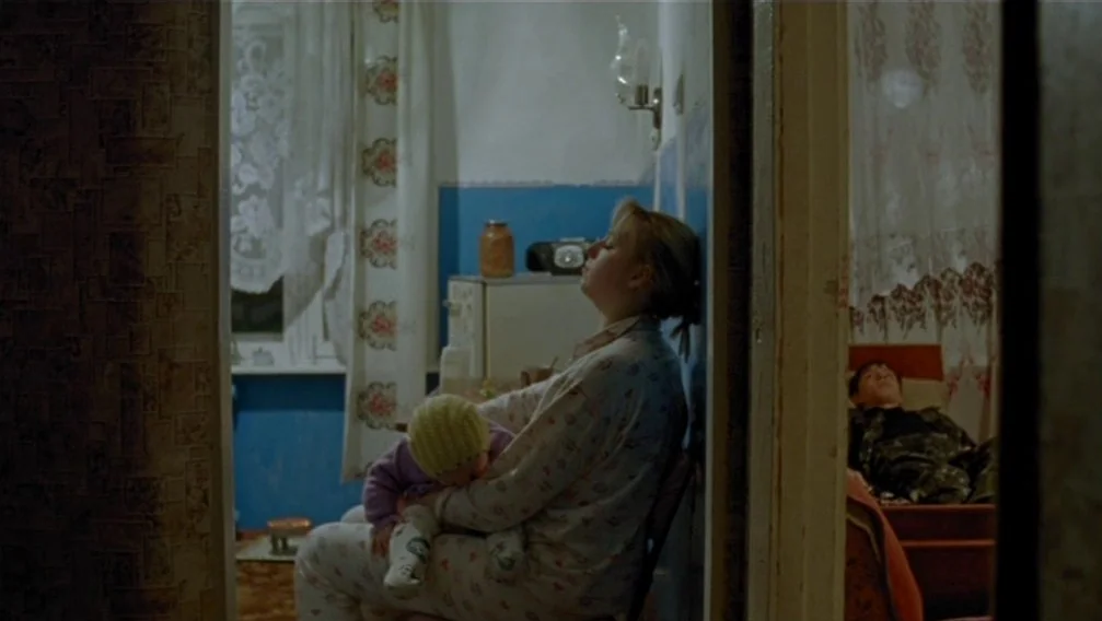 Import Export (2007) movie still: medium — A small apartment space with peeling wallpaper and blue painted sections near the floor. A woman sits in…