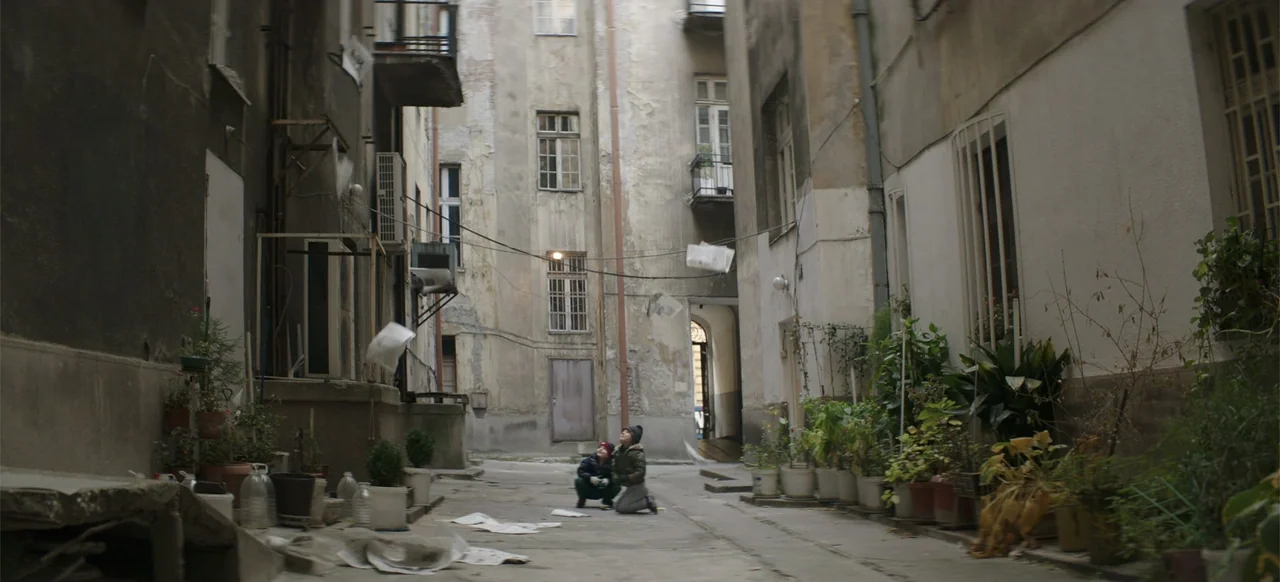 Morir un Poco (1967) movie still: wide — A weathered urban courtyard is surrounded by tall, aging apartment building walls with windows. Two young…