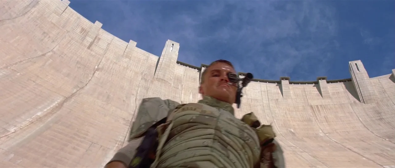 Universal Soldier (1992) movie still: medium — A massive concrete dam wall arcs across the background under a clear sky. A man in tactical armor…