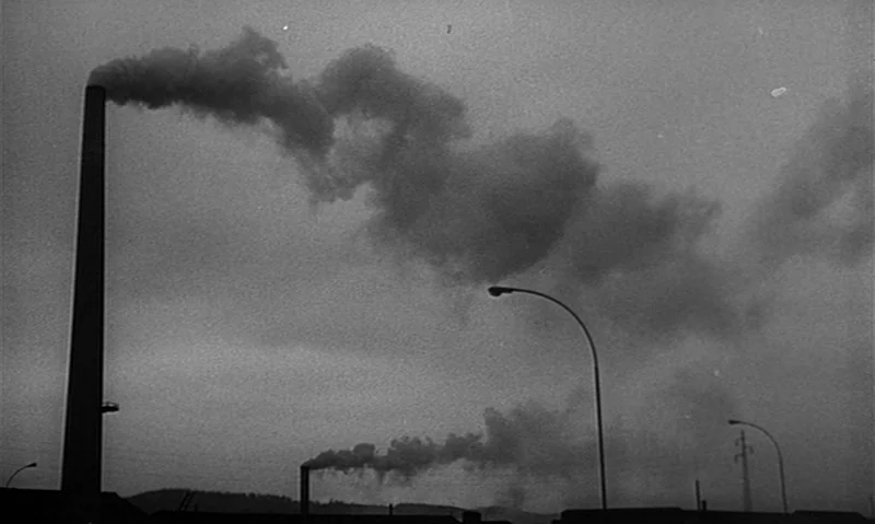 Man Is Not Bird (1965) movie still: establishing — Tall industrial chimneys release thick plumes of smoke against a grey, overcast sky. Two curved…