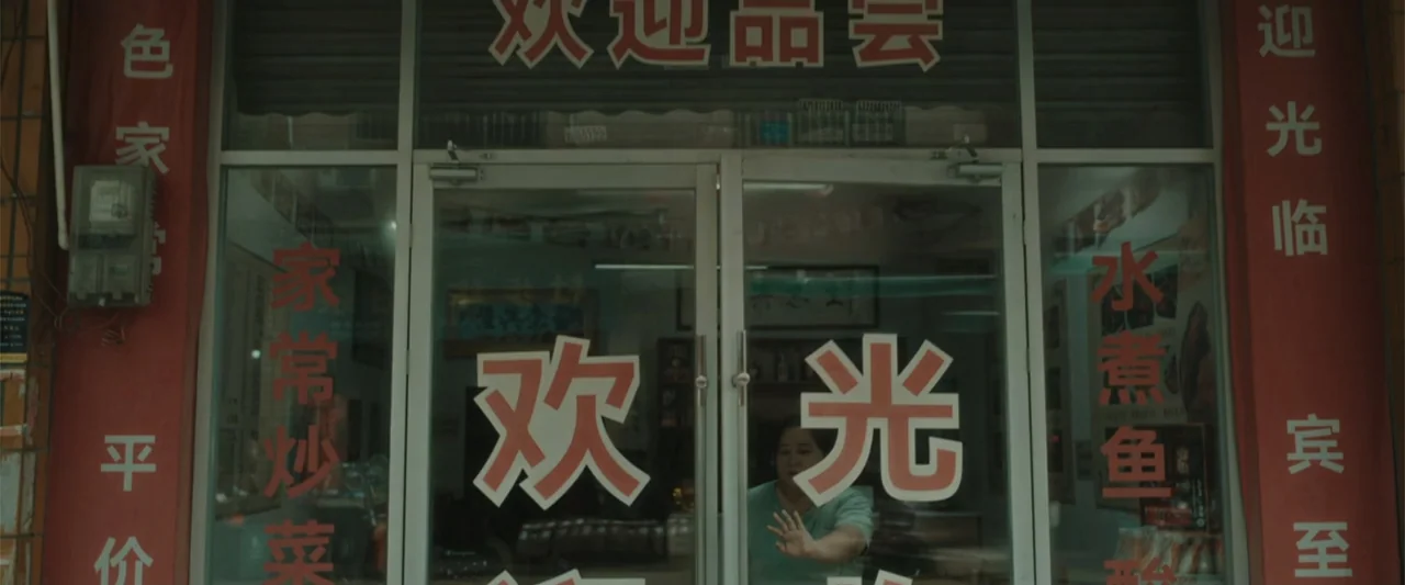 Yolo (2024) movie still: wide — A storefront with red vertical banners and large Chinese characters on the glass door. A middle-aged woman stands…