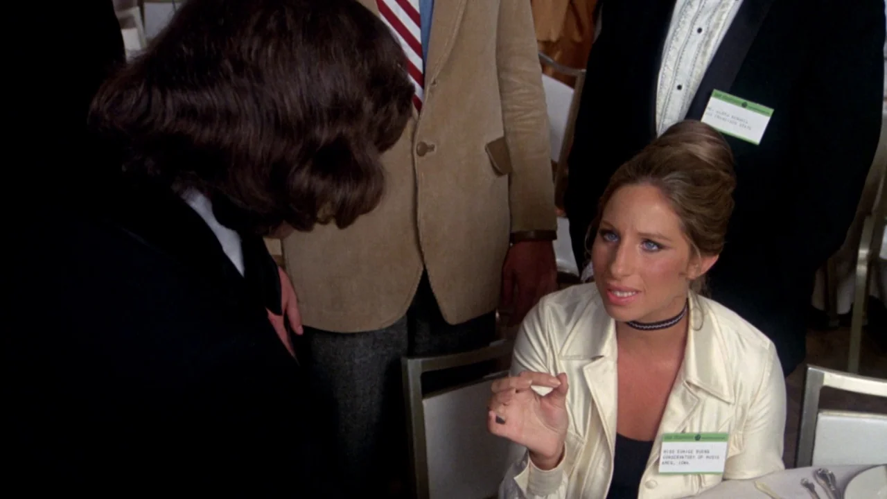 What’s Up, Doc? (1972) movie still: medium — A woman in a cream jacket sits at a table during a conference event. She gestures with her hand while…