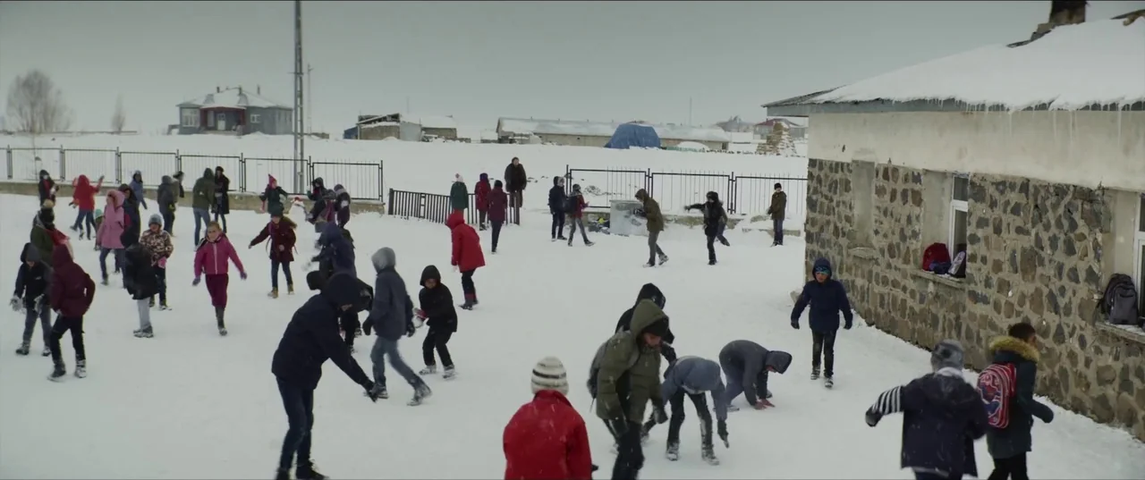 About Dry Grasses (2023) movie still: wide — A snowy outdoor schoolyard with a low stone building on the right side. Numerous children in winter…