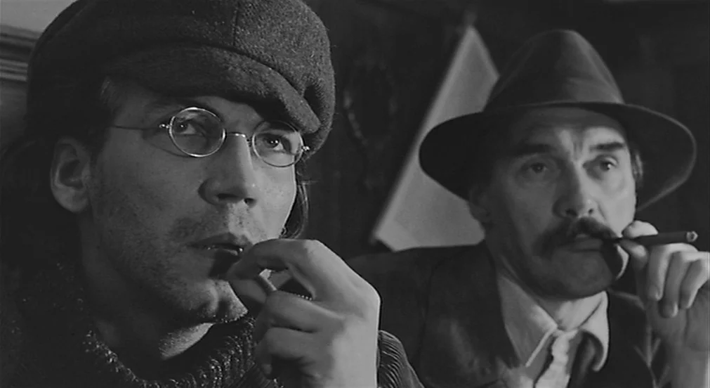 Kafka (1991) movie still: medium — Two men sit in a dimly lit, shadowed space that resembles a tavern setting. They both hold cigars near their…